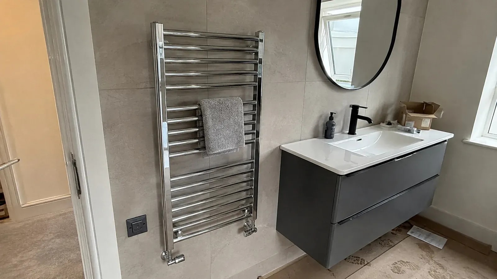 A chrome towel-warmer radiator beside a grey bathroom vanity with a white basin and oval mirror.