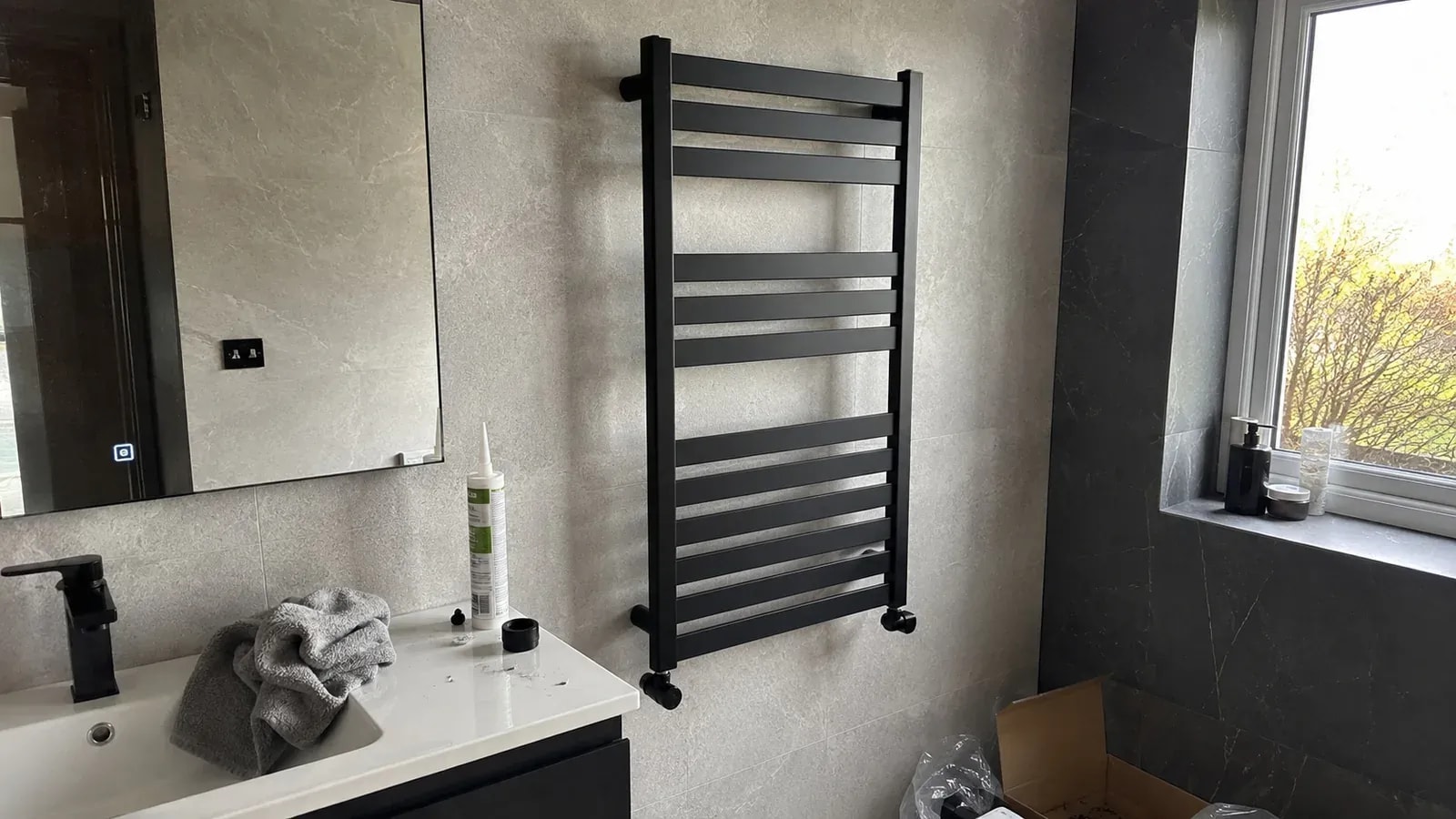 A flat black towel-warmer radiator above a bathroom vanity, with a silicone tube and installation tools on the counter.