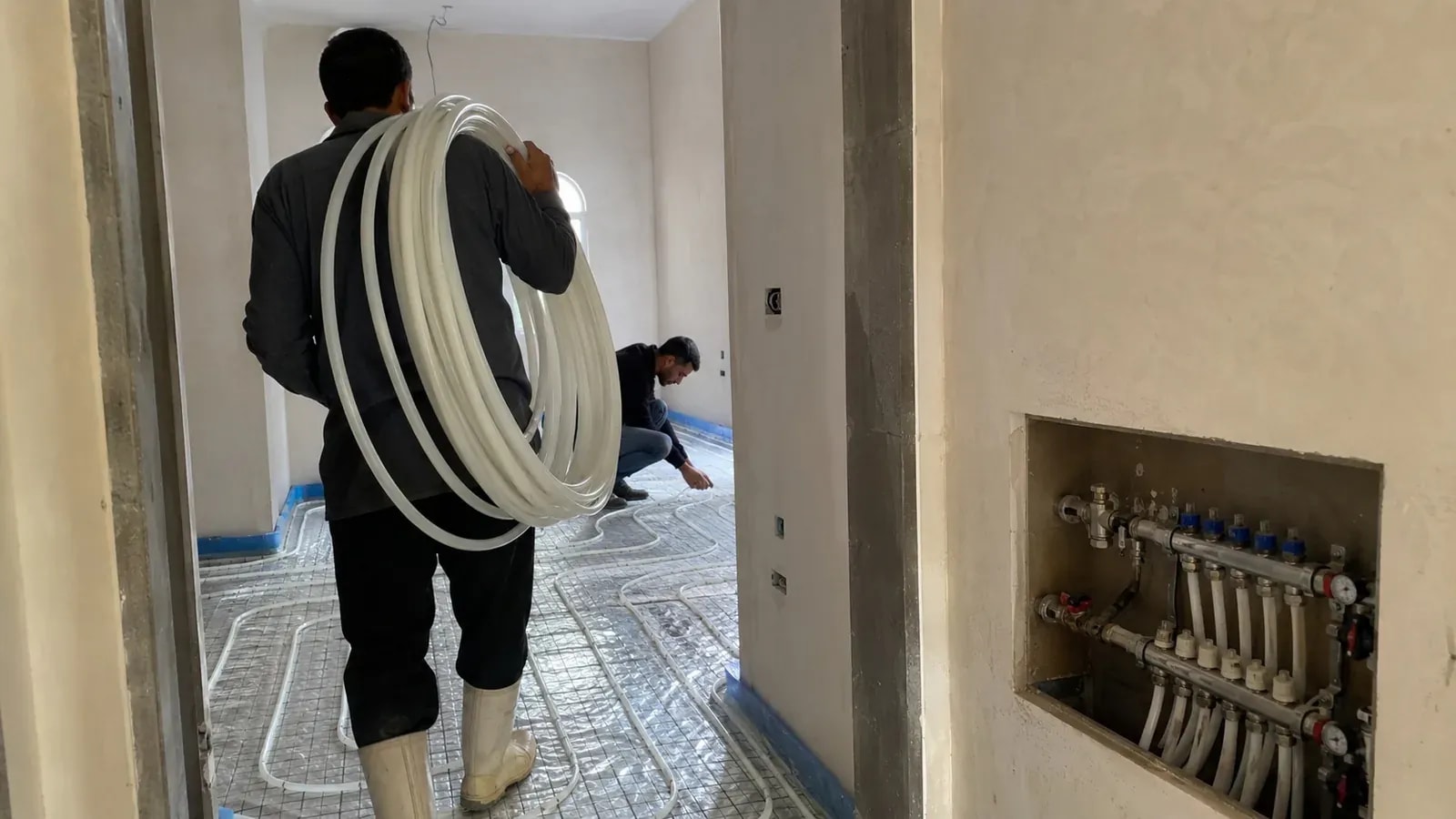 فنّي شايل لفة مواسير بيضا على كتفه وزميله بيركّب على أرضية التدفئة الأرضية جنب لوحة موزّع في الحيط